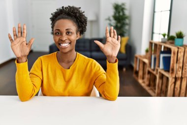 Young african american woman wearing casual clothes sitting on the table at home showing and pointing up with fingers number ten while smiling confident and happy. 