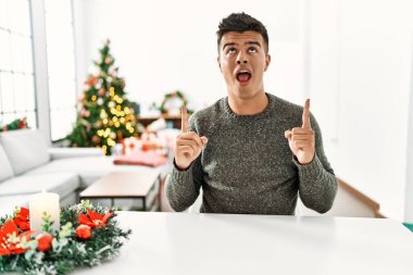 Noel ağacının yanında masada oturan İspanyol genç adam şaşırmış ve şaşırmış bir şekilde yukarı bakıyor ve parmaklarıyla işaret ediyor ve kollarını kaldırıyordu.. 