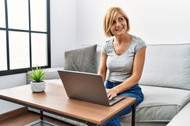 Orta yaşlı sarışın kadın evde dizüstü bilgisayar kullanarak gülümsüyor.