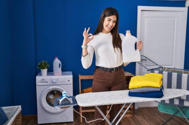 Ütüledikten sonra katlanmış çamaşırları olan genç esmer kadın parmaklarıyla imza atıp gülümseyerek mükemmel bir sembol oluşturuyor. 