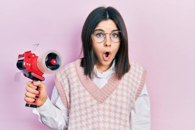 Young brunette woman holding packing tape scared and amazed with open mouth for surprise, disbelief face 