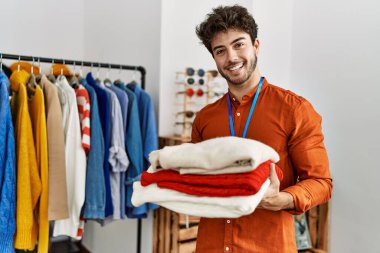 Genç İspanyol esnaf gülümseyerek giyim mağazasında bir yığın süveter tutuyor..