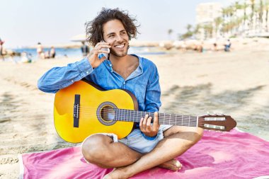Gitar çalan genç İspanyol adam kumsalda kumların üzerinde oturmuş akıllı telefonla konuşuyor..