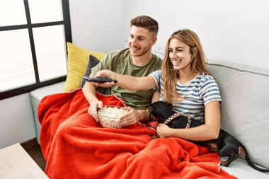 Genç İspanyol çift gülümsüyor. Mutlu bir şekilde kanepede oturmuş, evde köpekleriyle film izliyorlar..