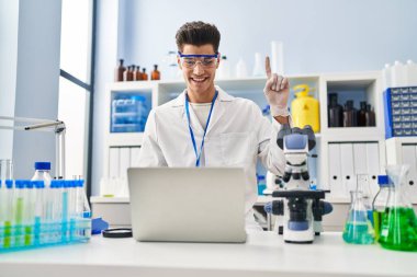 Young hispanic man working at scientist laboratory doing video call smiling with an idea or question pointing finger with happy face, number one 
