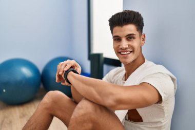Genç İspanyol adam spor merkezinde saatle yerde oturuyor.