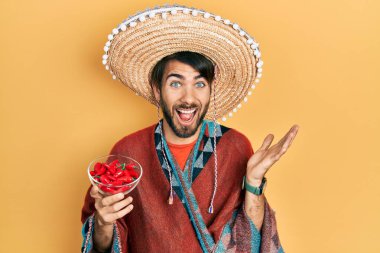 Young hispanic man wearing mexican hat holding chili celebrating victory with happy smile and winner expression with raised hands 