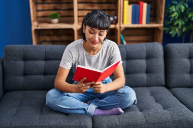 Evdeki kanepede kitap okuyan genç bir kadın.