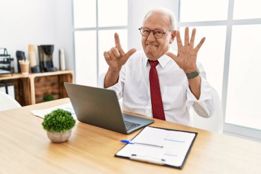 Senior man working at the office using computer laptop showing and pointing up with fingers number seven while smiling confident and happy. 