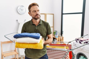 Middle age man holding folded laundry after ironing smiling looking to the side and staring away thinking. 