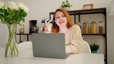 Genç kızıl saçlı kadın evde akıllı telefondan laptopla sesli mesaj dinliyor.