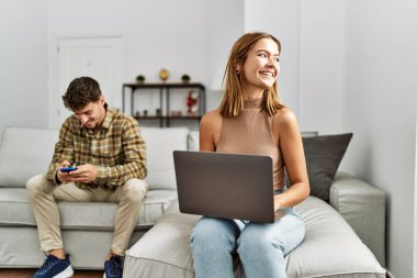 Genç İspanyol çift dizüstü bilgisayar ve akıllı telefon kullanarak mutlu bir şekilde gülümsüyor. Evdeki kanepede oturuyor..