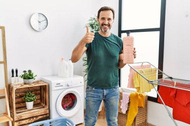 Middle age man with beard doing laundry holding detergent bottle smiling happy and positive, thumb up doing excellent and approval sign 