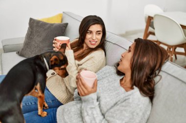 İki kadın kanepede oturmuş köpeğiyle kahve içiyor.