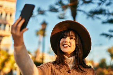 Kışlık şapkalı esmer kadın parkta akıllı telefonuyla selfie çekiyor.