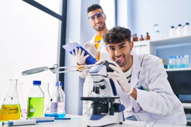 Mikroskop kullanan iki bilim adamı laboratuarda çalışan panoya yazı yazıyorlar.