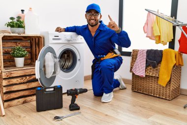 Young indian technician working on washing machine smiling amazed and surprised and pointing up with fingers and raised arms. 