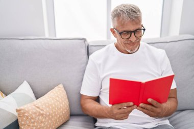 Orta yaşlı, kır saçlı, kitap okuyan adam evdeki kanepede oturuyor.
