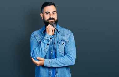 Sakallı, sıradan kot ceketli İspanyol bir adam kameraya kollarını kavuşturup çenesine dokunarak gülümsüyor. Olumlu düşünmek. 