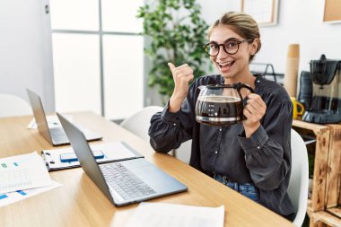 Ofiste çalışan genç sarışın kadın elinde kahve makinesiyle baş parmağını kaldırıyor ve ağzı açık gülümsüyor. 