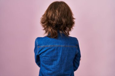 Orta yaşlı bir kadın pembe arka planda dikiliyor. Arkaya bakıyor ve kollarını kavuşturuyor. 
