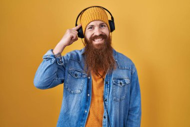 Caucasian man with long beard listening to music using headphones smiling pointing to head with one finger, great idea or thought, good memory 
