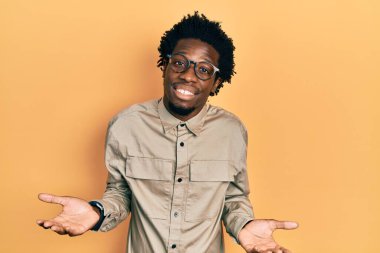 Sıradan kıyafetler giyen, gözlüklü, kafası karışmış, elleri ve kolları havada genç Afro-Amerikan bir adam. şüphe kavramı. 