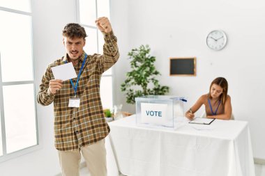 Young handsome man voting putting envelop in ballot box annoyed and frustrated shouting with anger, yelling crazy with anger and hand raised 