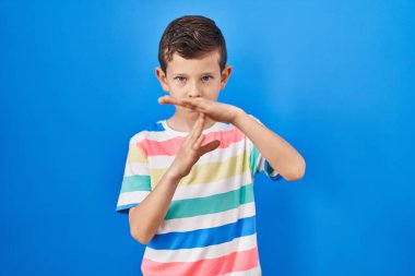 Mavi arka planda dikilen genç beyaz çocuk elleriyle jest yapıyor, hayal kırıklığına uğramış ve ciddi bir yüzü var. 