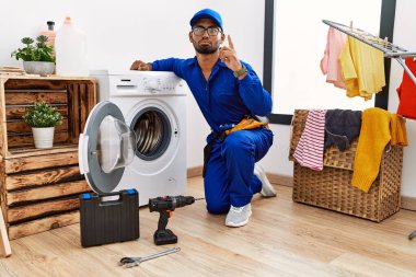 Young indian technician working on washing machine pointing up looking sad and upset, indicating direction with fingers, unhappy and depressed. 