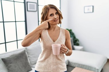Orta yaşlı bir kadın evde kahve içerken parmakları birbirine dönük, neşeli bir şekilde gülümsüyor. güzellik konsepti 