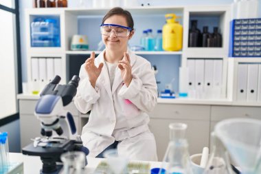 Hispanic girl with down syndrome working at scientist laboratory gesturing finger crossed smiling with hope and eyes closed. luck and superstitious concept. 