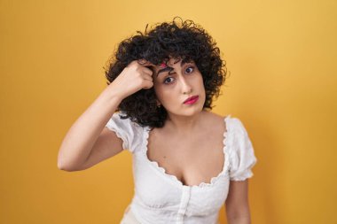 Young brunette woman with curly hair standing over yellow background pointing unhappy to pimple on forehead, ugly infection of blackhead. acne and skin problem 