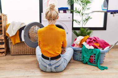 Young blonde woman doing laundry sitting by washing machine standing backwards looking away with crossed arms 