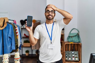 Young hispanic man with beard working as manager at retail boutique holding smartphone stressed and frustrated with hand on head, surprised and angry face 