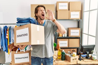 Handsome middle age man holding donations box for charity at volunteer stand shouting and screaming loud to side with hand on mouth. communication concept. 