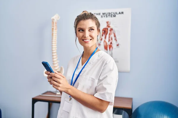 Young Blonde Woman Wearing Physiotherapist Uniform Using Smartphone Clinic — стоковое фото