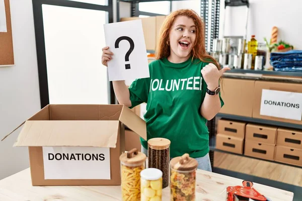 年轻的红头发女人穿着志愿者的T恤 手里拿着问号 手指指向旁边 微笑着张开嘴 — 图库照片