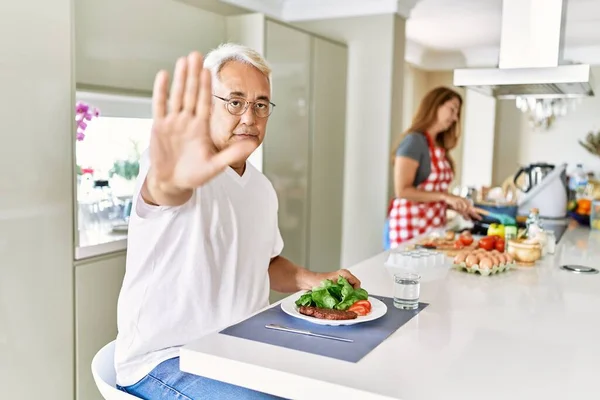 Middle age hispanic couple eating healthy meal at home with open hand doing stop sign with serious and confident expression, defense gesture