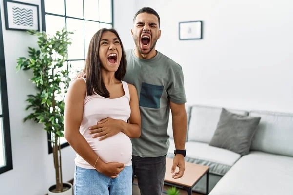 Young interracial couple expecting a baby, touching pregnant belly angry and mad screaming frustrated and furious, shouting with anger. rage and aggressive concept.