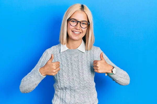 Beautiful blonde woman wearing glasses success sign doing positive gesture with hand, thumbs up smiling and happy. cheerful expression and winner gesture.