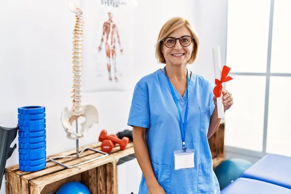 Middle age blonde woman wearing physiotherapist uniform holding diploma at clinic