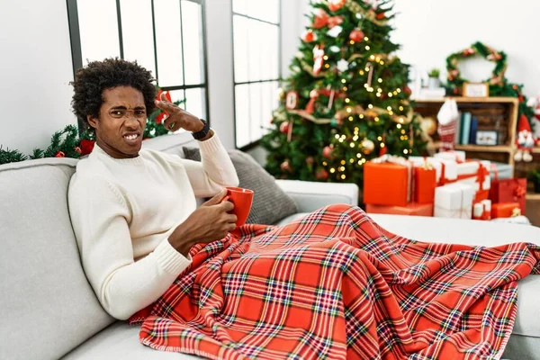 若いアフリカ系アメリカ人男性座っているソファの上にコーヒーを飲みながらクリスマスツリーを撮影し 銃のように頭に手や指を指して自分自身を殺す 自殺ジェスチャー — ストック写真
