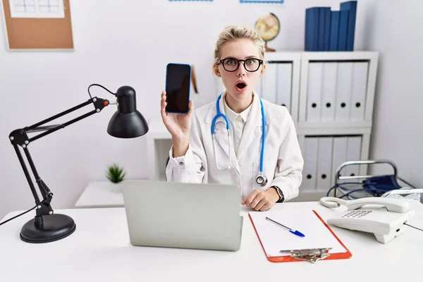 Young Doctor Woman Holding Smartphone Showing Blank Screen Scared Amazed — Fotografia de Stock