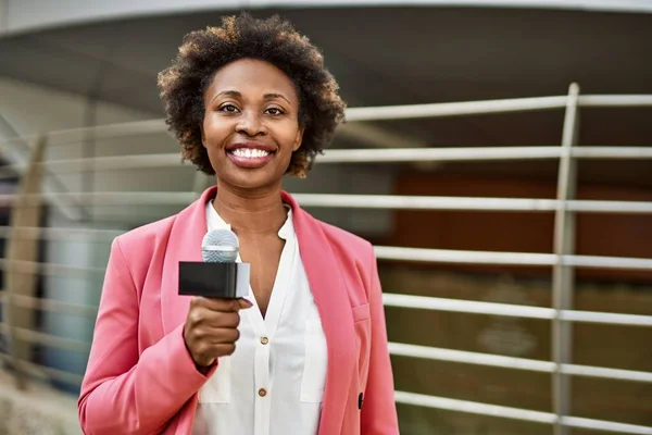 Young african american woman journalist holding reporter microphone speaking and smiling to the camera for television news