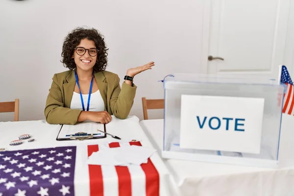 Young hispanic woman at political election sitting by ballot smiling cheerful presenting and pointing with palm of hand looking at the camera.