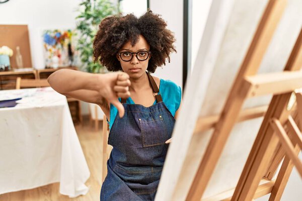 Beautiful african american woman with afro hair painting canvas at art studio with angry face, negative sign showing dislike with thumbs down, rejection concept 