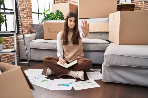 Young brunette woman moving to a new home doing finances with open hand doing stop sign with serious and confident expression, defense gesture