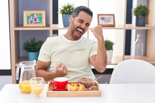 Hispanic man with beard eating breakfast celebrating surprised and amazed for success with arms raised and eyes closed