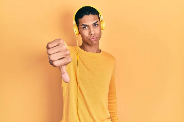 年轻的非洲裔美国人 一边用耳机听音乐 一边看起来很不高兴 很生气 用拇指低下的手势表示拒绝和消极 不好的表达方式 — 图库照片